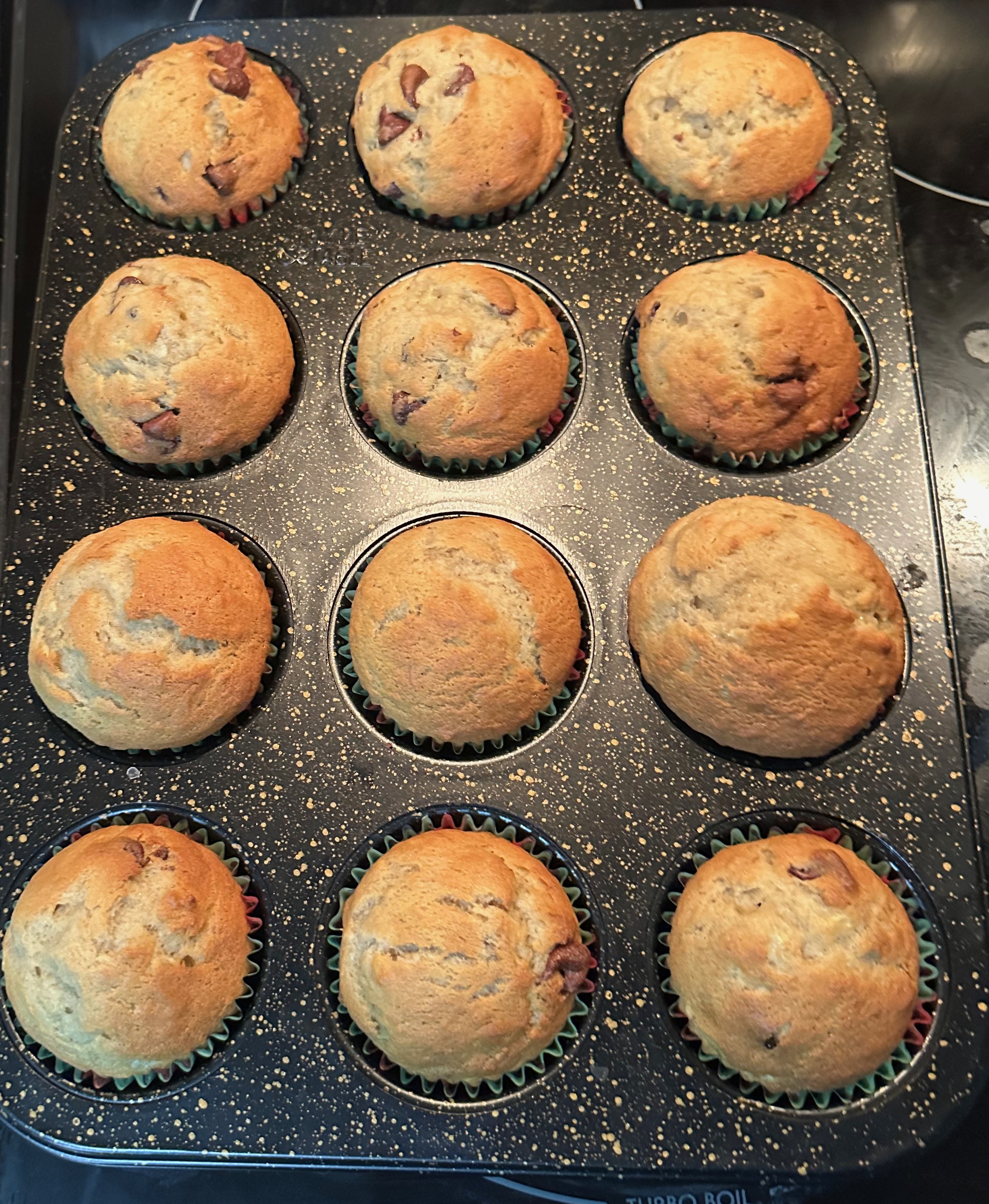 Twelve golden-brown banana chocolate chip muffins cooling in a dark muffin tin