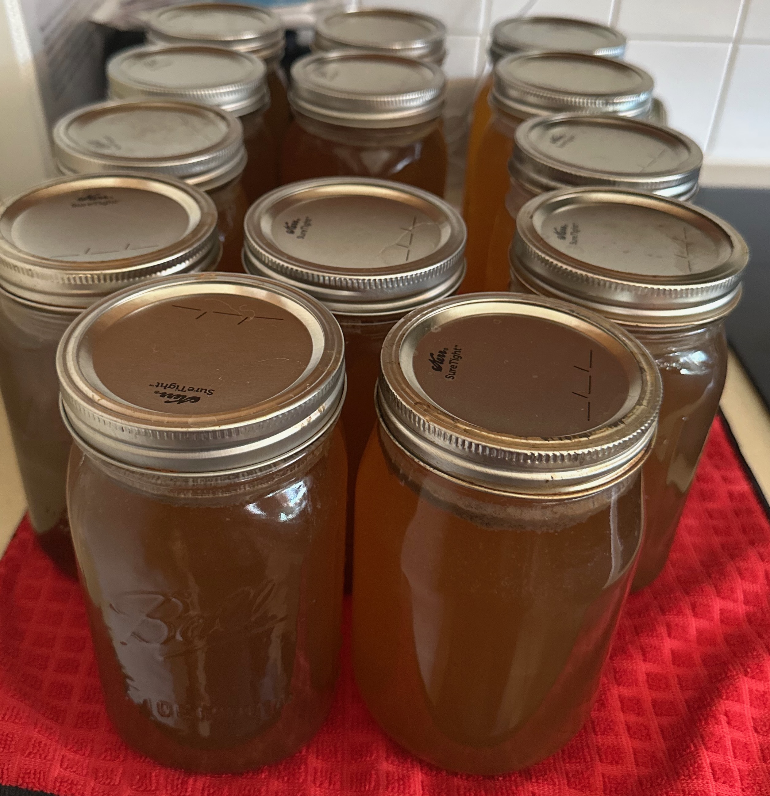 Twelve sealed Ball mason jars of amber bone broth cooling on a red kitchen cloth