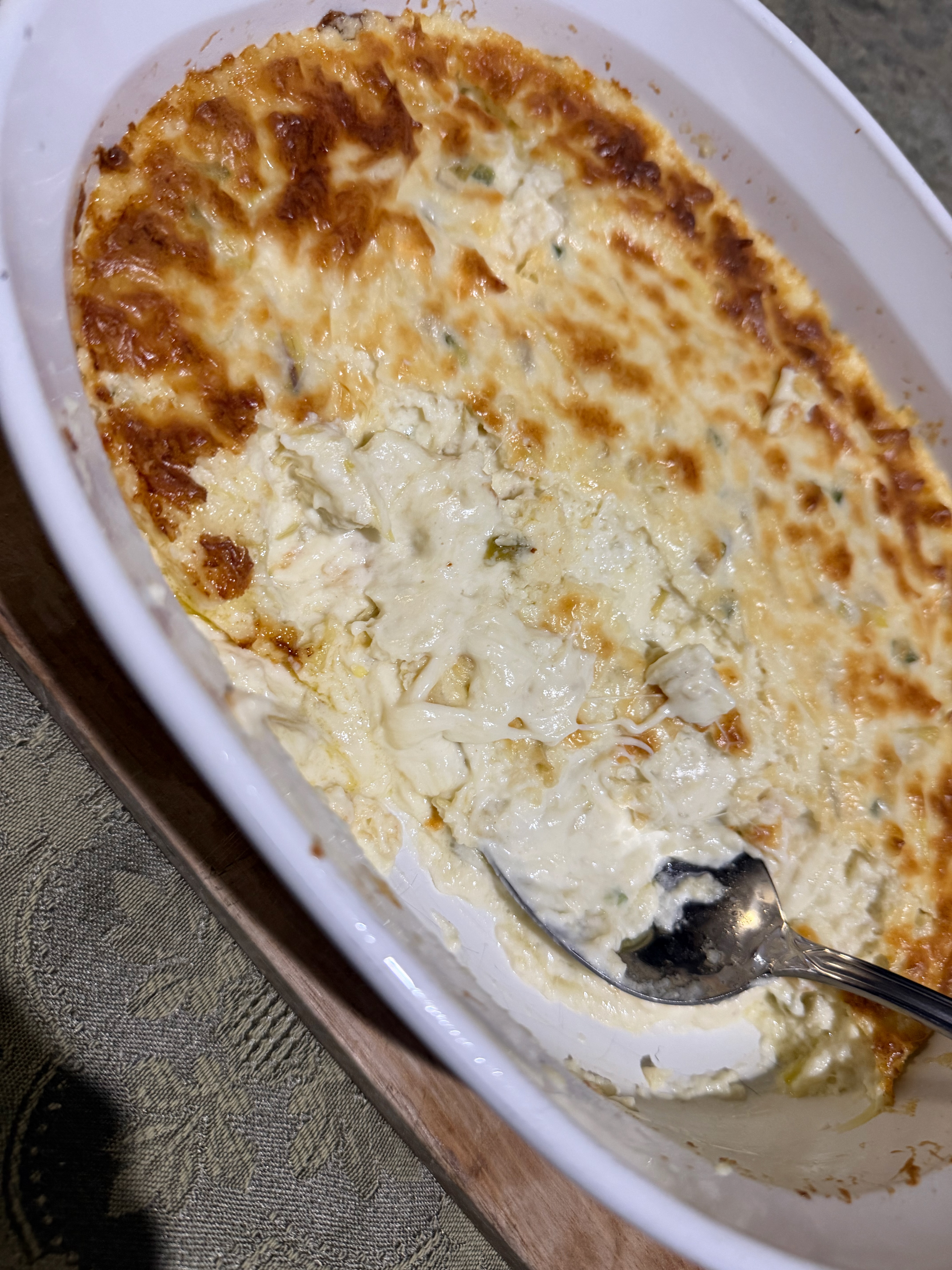 A white oval baking dish of hot artichoke dip with a golden-brown bubbling cheese top, with a serving spoon