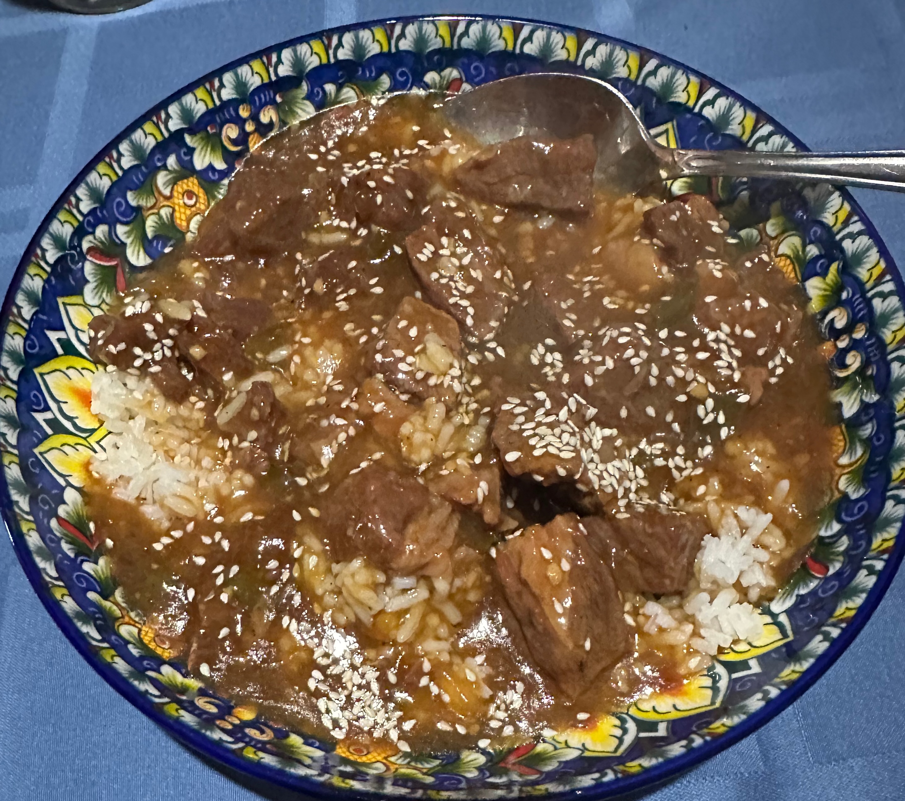 A decorative folk-art bowl of Korean beef in rich dark sauce over white rice, topped with sesame seeds