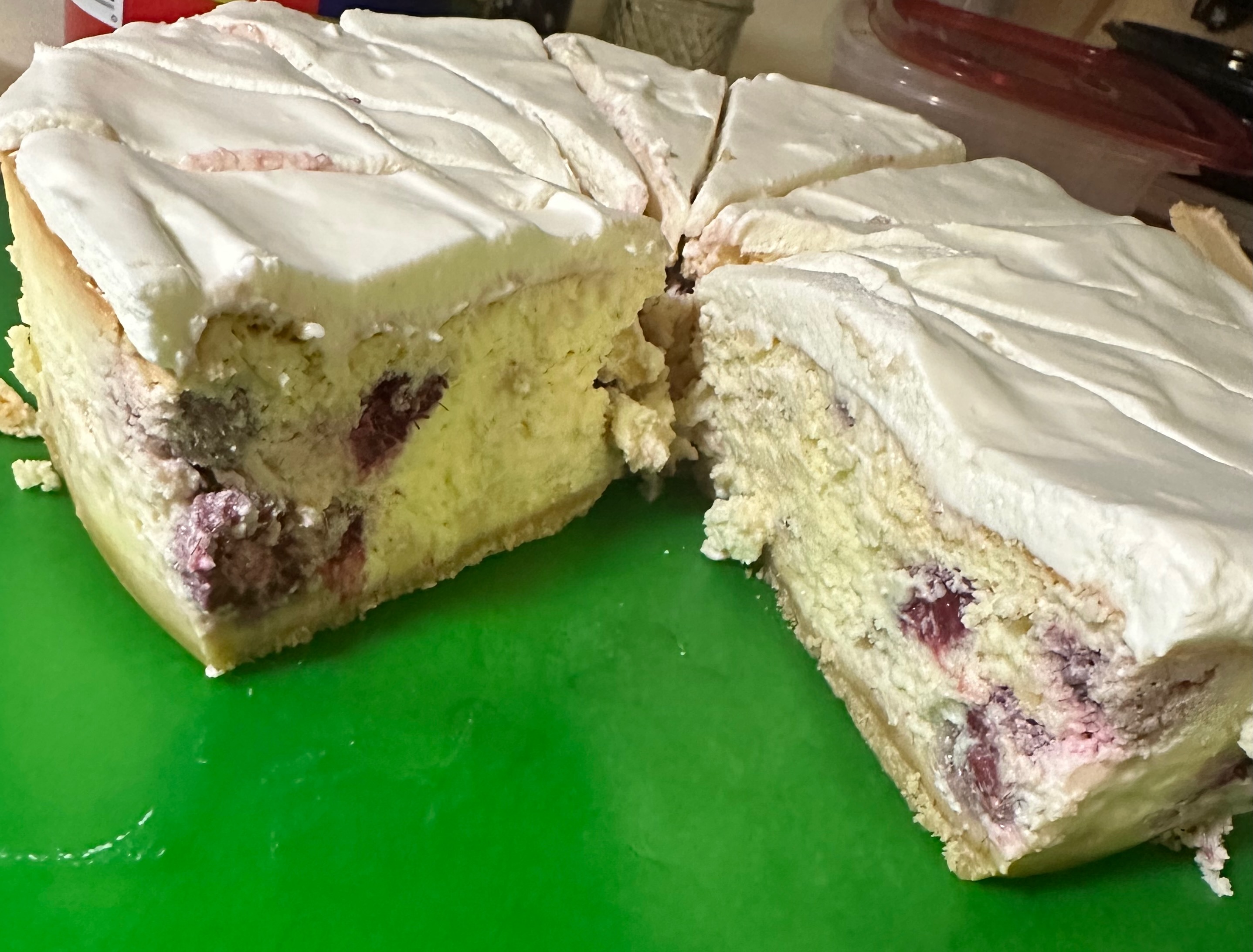 Sliced cheesecake with fluffy white cream topping and visible raspberry pieces in the pale yellow filling, on a green cutting board