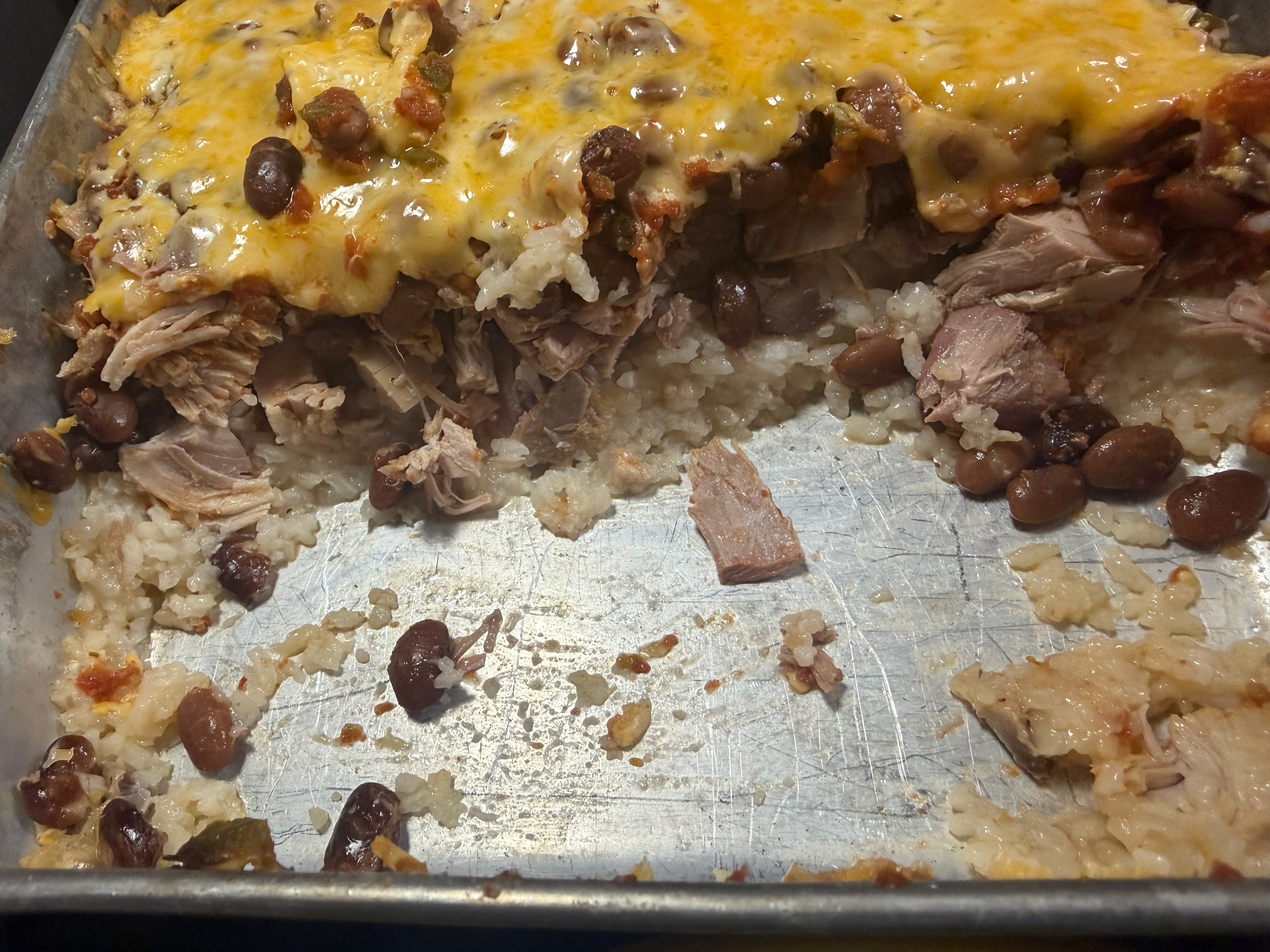 A 9×13 pan of Mexican casserole with pulled pork, black beans, and rice topped with melted shredded cheese, partially served