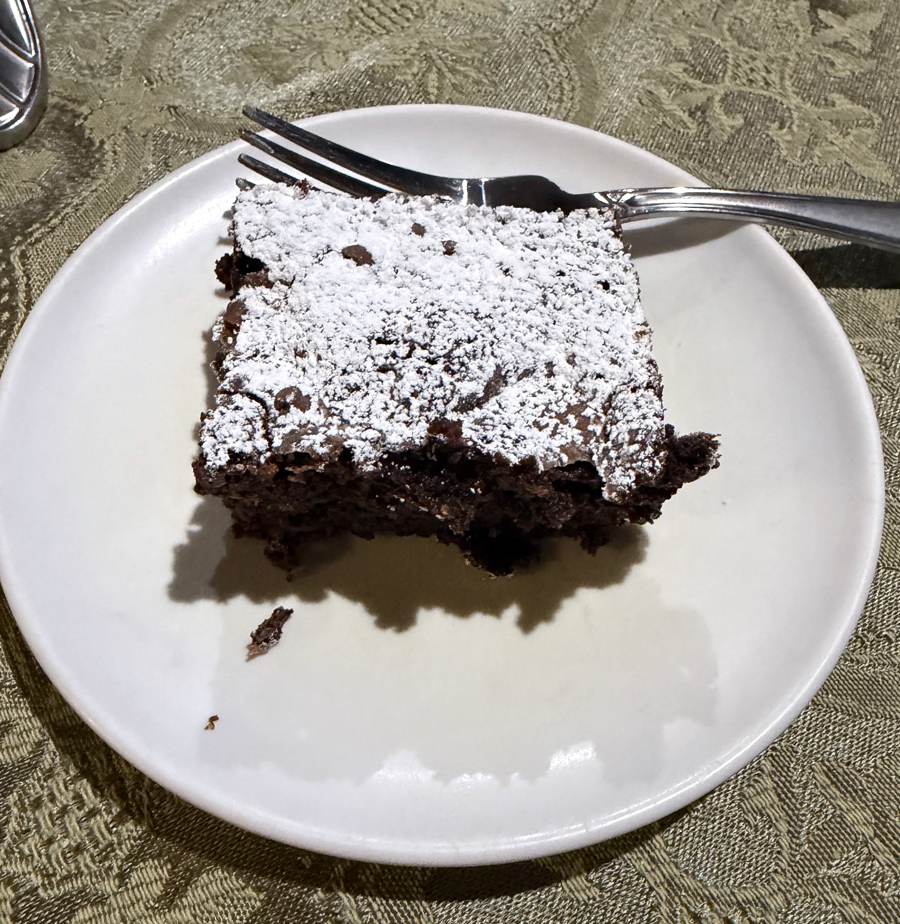 A thick fudgy brownie square dusted with powdered sugar on a white plate with a fork